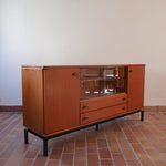 Enfilade vintage en teck années 60, deux portes pleines encadrant une vitrine centrale à portes coulissantes en verre, deux tiroirs, poignées navette métal, piétement métal laqué noir
