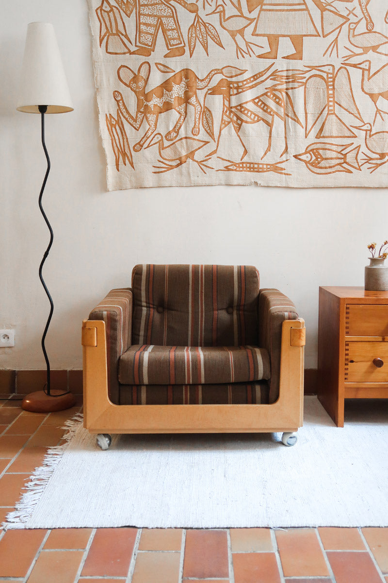 Fauteuil moderniste années 70 en bois massif clair sur roulettes avec coussins rayés bruns et terracotta.