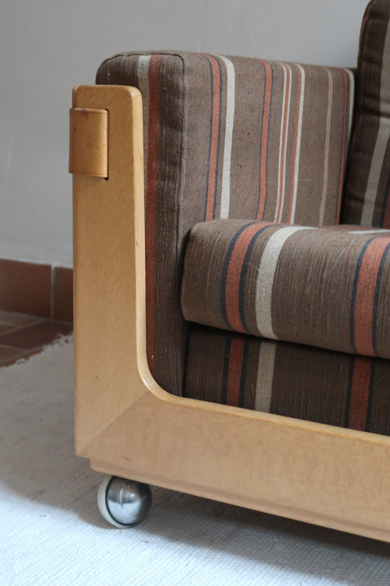 Fauteuil moderniste années 70 en bois massif clair sur roulettes avec coussins rayés bruns et terracotta.