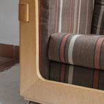 Fauteuil moderniste années 70 en bois massif clair sur roulettes avec coussins rayés bruns et terracotta.