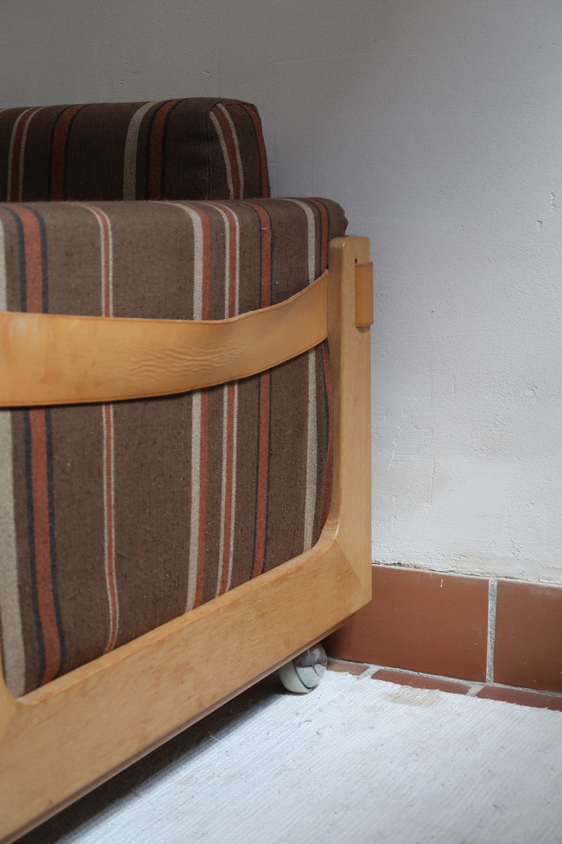 Fauteuil moderniste années 70 en bois massif clair sur roulettes avec coussins rayés bruns et terracotta.