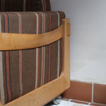 Fauteuil moderniste années 70 en bois massif clair sur roulettes avec coussins rayés bruns et terracotta.