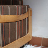 Fauteuil moderniste années 70 en bois massif clair sur roulettes avec coussins rayés bruns et terracotta.