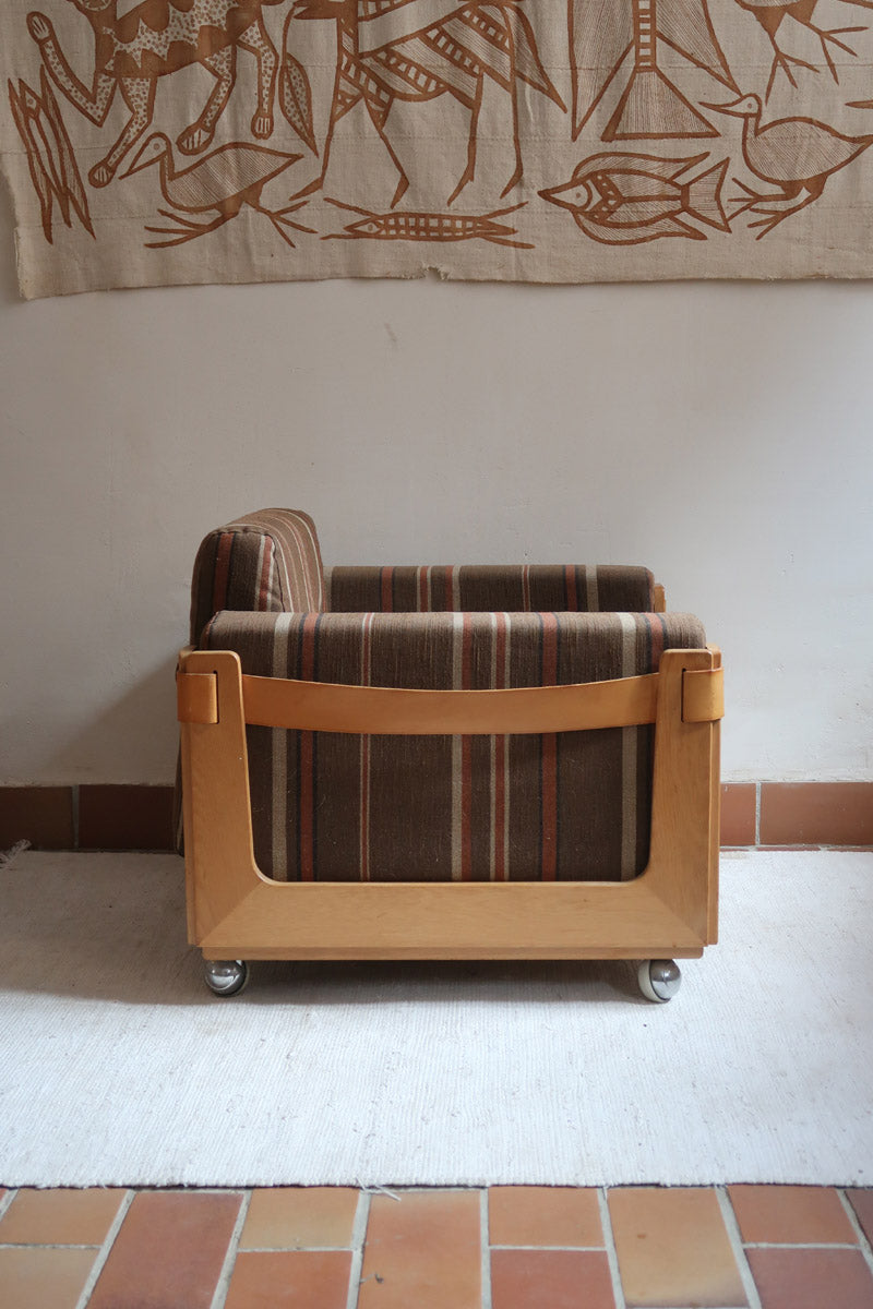 Fauteuil moderniste années 70 en bois massif clair sur roulettes avec coussins rayés bruns et terracotta.