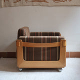Fauteuil moderniste années 70 en bois massif clair sur roulettes avec coussins rayés bruns et terracotta.