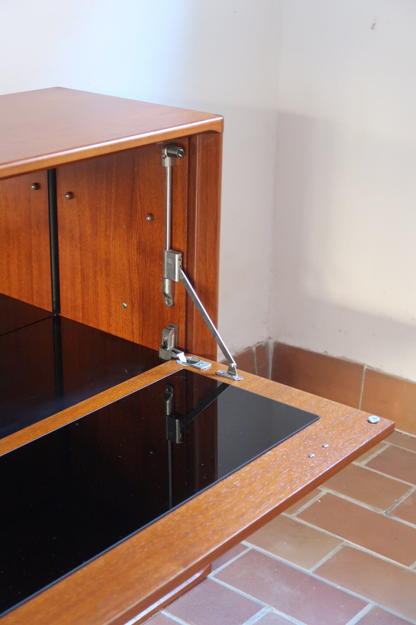 Meuble bar vintage en bois de teck, années 90, abattant sur deux compas chromés découvrant un intérieur doublé de verre noir, poignée sculptée dans la masse, niche basse ouverte sur toute la largeur