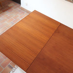 Table à manger scandinave en teck avec deux rallonges intégrées, design années 60, photographiée dans un intérieur lumineux.