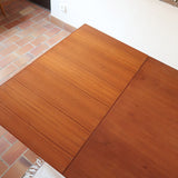 Table à manger scandinave en teck avec deux rallonges intégrées, design années 60, photographiée dans un intérieur lumineux.