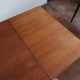 Table à manger scandinave en teck avec deux rallonges intégrées, design années 60, photographiée dans un intérieur lumineux.