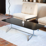 Table basse design années 70 avec plateau noir rectangulaire et piétement en acier brossé en forme de patins.