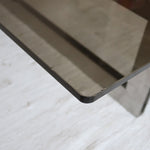 Table basse vintage en verre fumé, années 70, grand plateau rectangulaire à angles arrondis, piétement en inox brossé composé de deux cadres en U réunis par une croix centrale