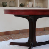 Table à manger ovale en teck avec piètement central sculptural en bois foncé et plateau à teinte rougeâtre, style scandinave vintage, présentée seule et en mise en situation.